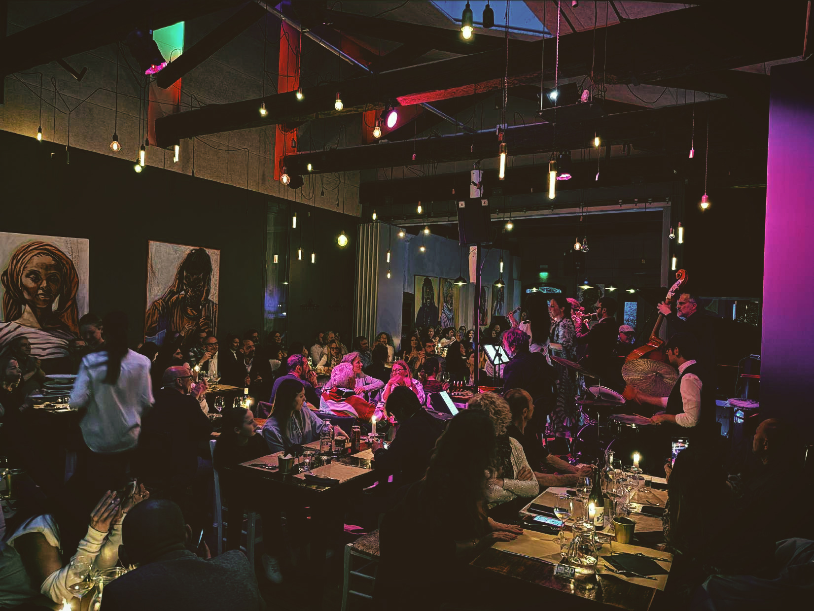 Restaurant filled with diners watching a live jazz band on a small stage; musicians play saxophone, upright bass and drums under warm hanging lights and colorful ambience.